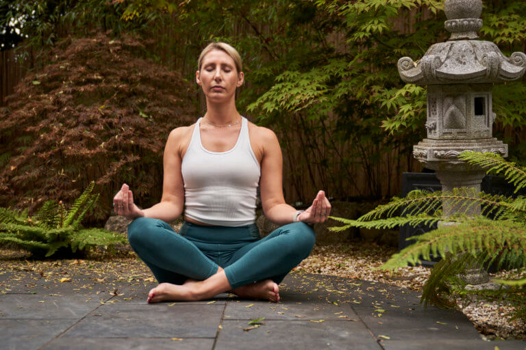 A female sitting cross-legged and meditating in a garden with a Thai statue surrounded by azaleas and ferns.