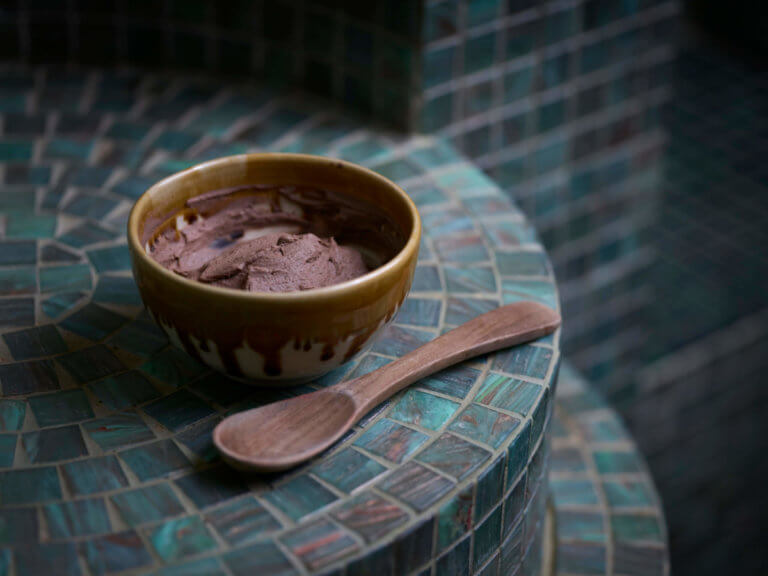A bowl of Rhassoul mud on the side of a green tiled steam room with a wooden spoon in the foreground.