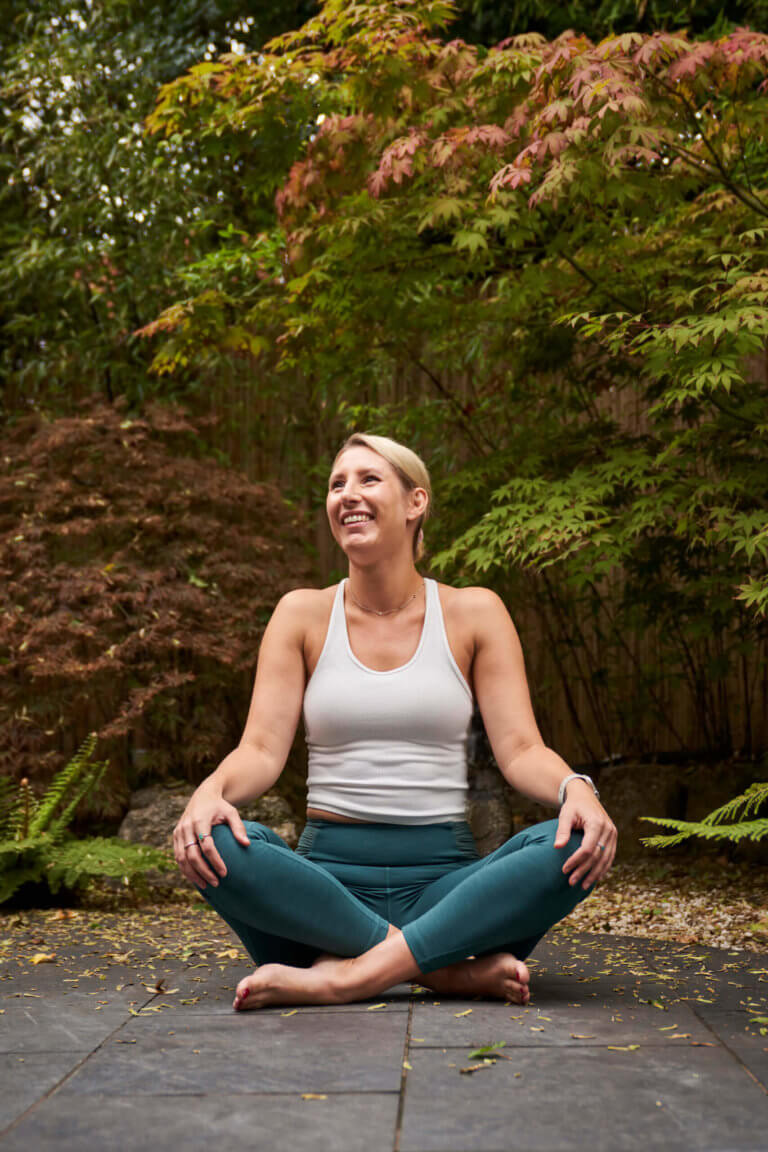 Woman sits in garden cross legged with hands on knees smiling