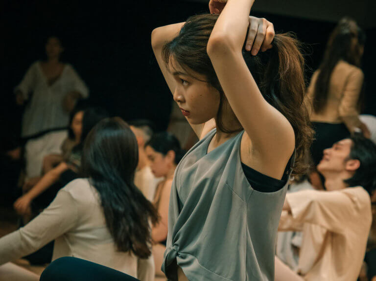 A female sitting cross-legged in a dance studio with a group of people. She is tying her long brown hair back.