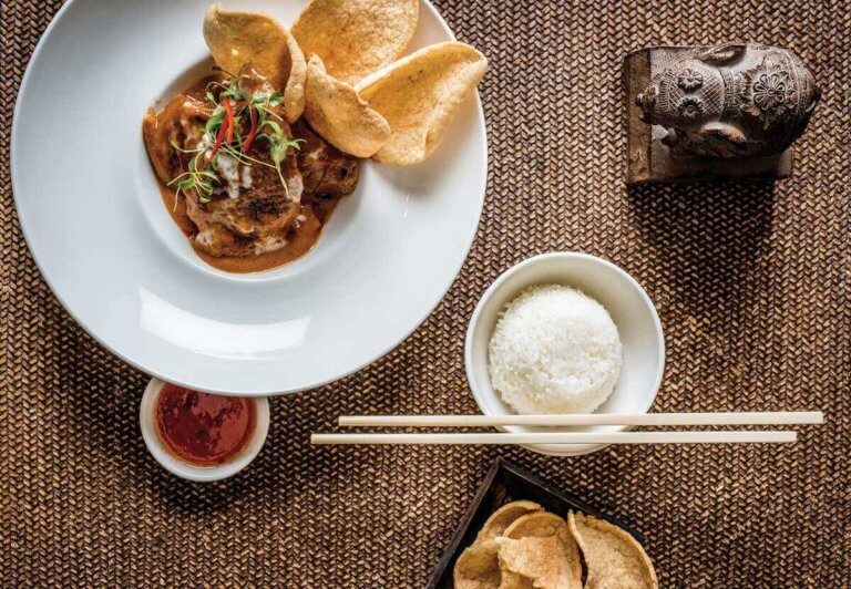 A flat-lay of a Thai duck dish in a white bowl with Thai crackers on the side . There is a small bowl of sweet chili sauce and a bowl of jasmine rice next to it.