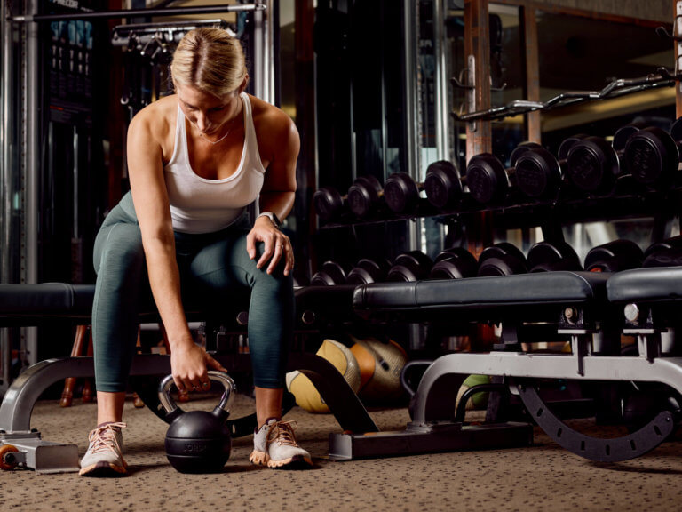 Woman sits on bench in gym and leans down to pick up a kettlebell with one hand