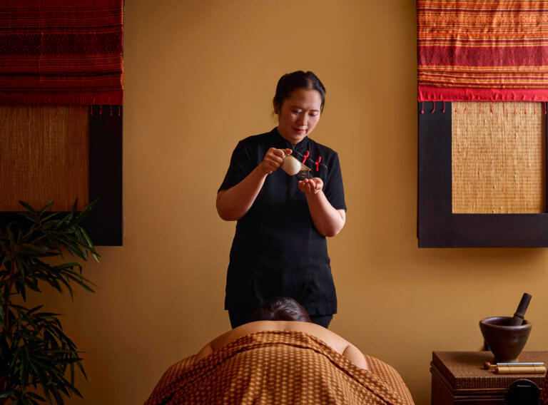 A spa therapist pouring oil from a small white jug into the palm of their hand. A female is lying on her front on a massage couch under Thai patterned fabrics.