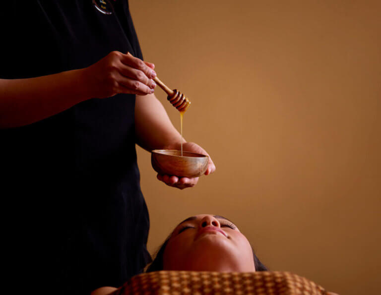 A spa therapist holding a honey dipper over a wooden bowl of manuka honey. Their spa client is lying down and relaxing.