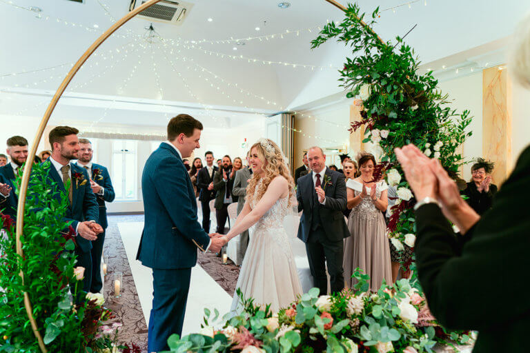 The bride and groom stand at the registrars table holding hand