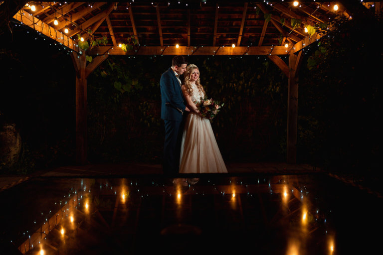 The married couple stand under fairy lights in the garden at night