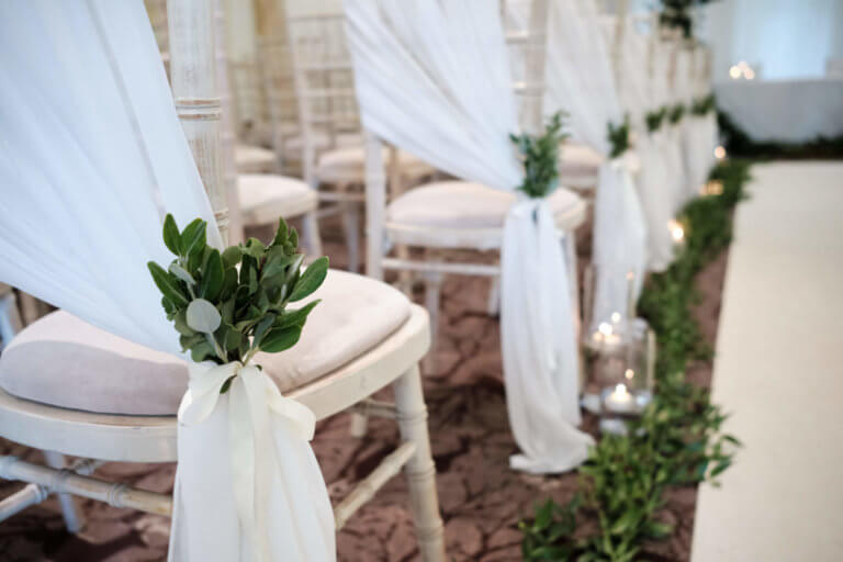 Wooden chairs with white sashes and green foliage at wedding ceremony