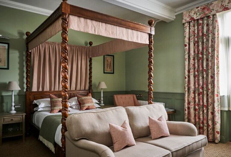 A traditional style hotel bedrooms with sage green walls and paneling, pink and cream floral curtains an oak fourposter bed and a beige couch.
