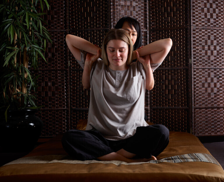 Thai therapist performs a traditional Thai massage with the recipient sitting cross legged and the therapist behind with her arms over her shoulders.