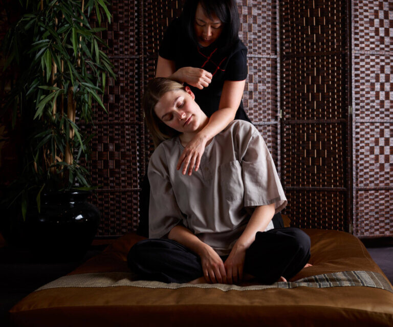 Thai therapists performs a traditional Thai massage with the recipient sitting cross legged and the therapist behind with her arms over her shoulders.