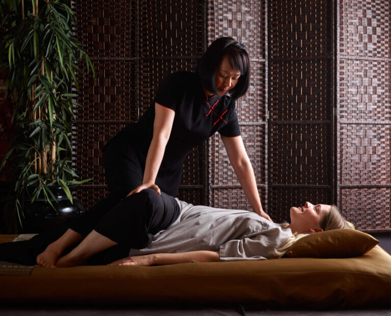Thai therapists performs a traditional Thai massage with the recipient lying down and the therapist leaning over her, bending one leg over the other