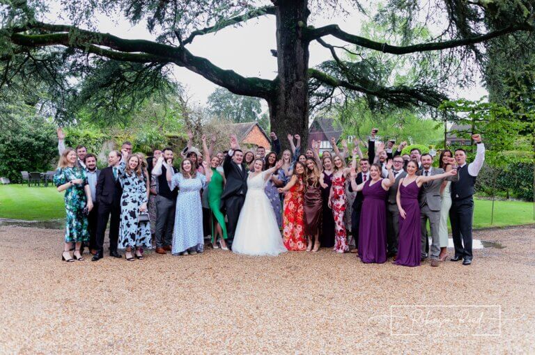 Whole wedding party cheers under tree on front lawn of Careys Manor Hotel & SenSpa wedding venue in The New Forest