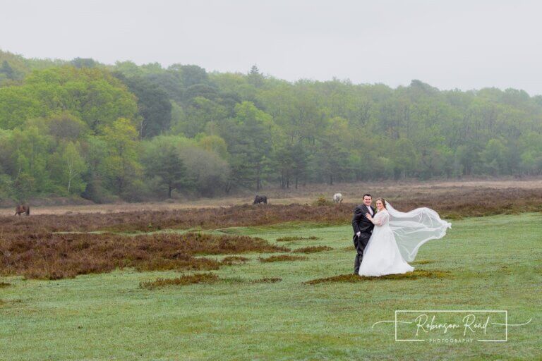 Bride and groom stand in windy field in The New Forest national park, with New Forest ponies in the background