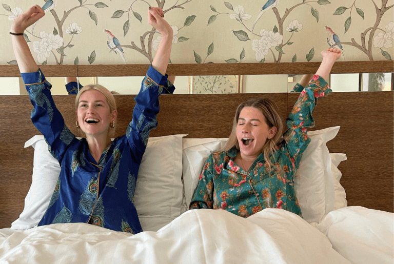 two friends waking up in bed after a great nights sleep at a hotel. they are both in white sheets and wearing silk-patterned pajamas.