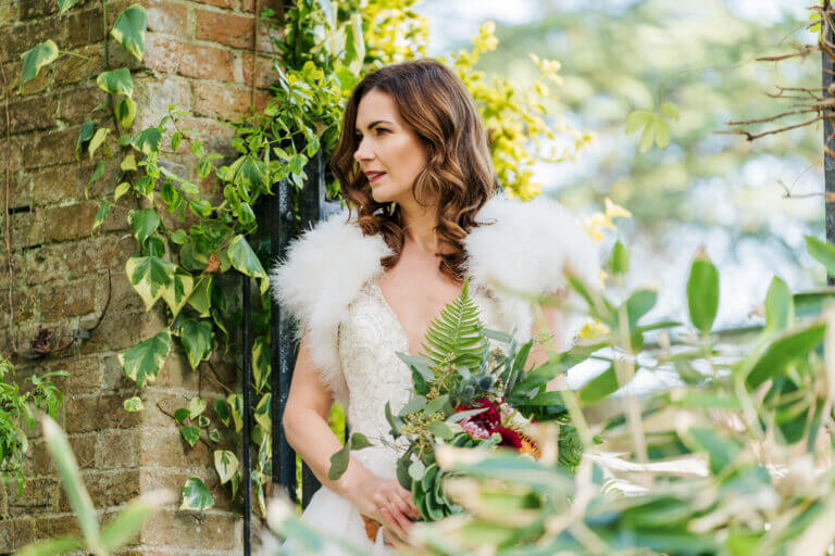 Bride walks through gardens in winter wedding dress with feather bolero carrying bouquet