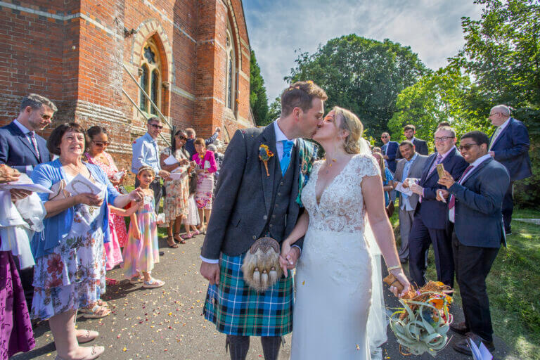 Bride and groom kiss outside church surrounded by wedding guests and flower petals on sunny day