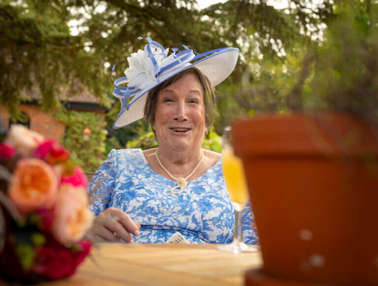Very happy wedding guest in blue floral dress and matching floral hat enjoys a drink on the lawn at Careys Manor Hotel wedding venue