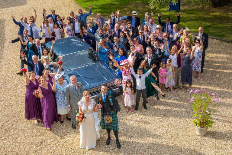 Birds eye shot of wedding party cheering, hands in the air, surrounding a classic car in the grounds of Careys Manor Hotel in Brockenhurst