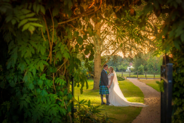 Bride and groom hold hands beneath large leafy green tree in Careys Manor Hotel grounds at dusk