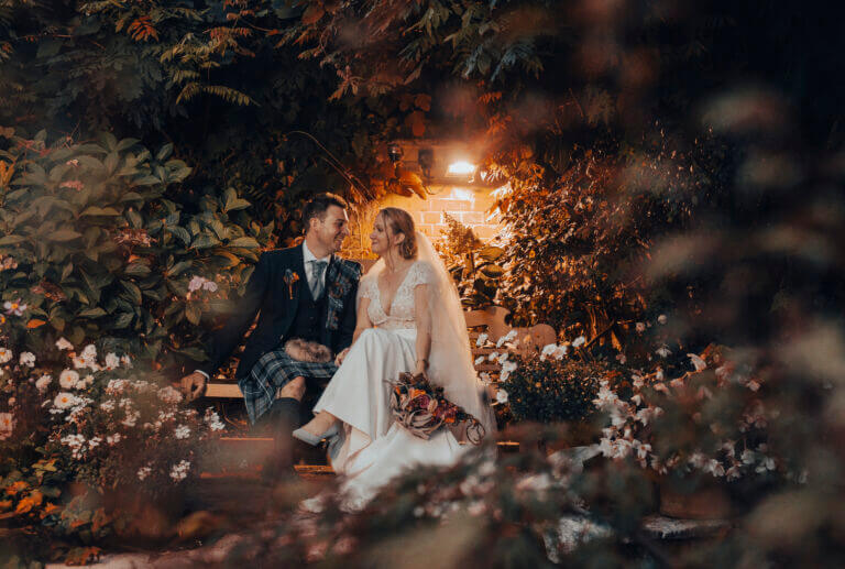 Bride and groom look lovingly at each other beneath trees whilst sitting on a bench in Careys Manor Hotel grounds at dark, with warm light above