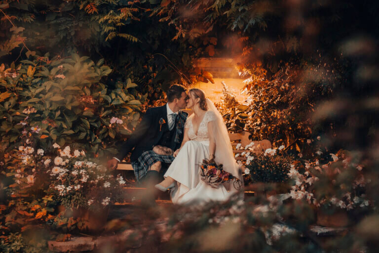 Bride and groom kiss beneath trees whilst sitting on a bench in Careys Manor Hotel grounds at dark, with warm light above