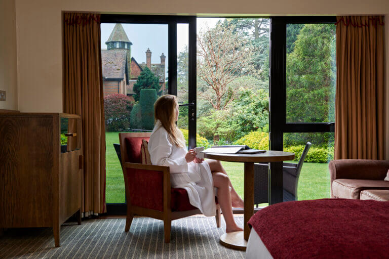Woman sits at table in bath robe looking out at gardens in Careys Manor Hotel garden room