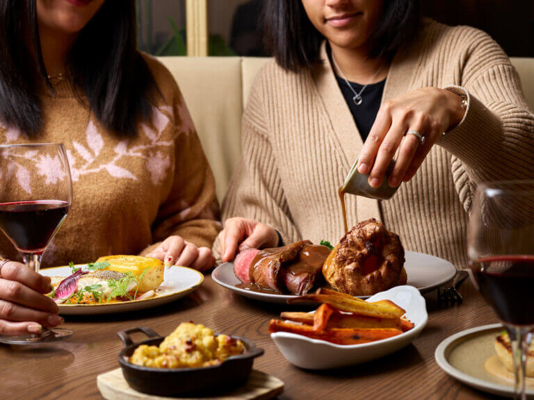 Two women enjoy Sunday lunch at Cambium restaurant in Brockenhurst as one pours gravy over beef dish and one holds a glass of red wine