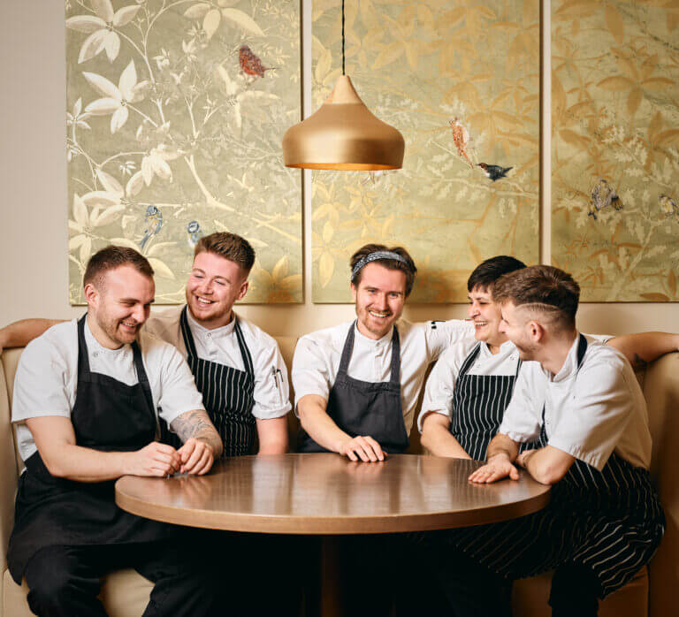 Group shot of happy chefs at Careys Manor hotel sitting at table in Cambium restaurant in their work uniforms with black aprons 