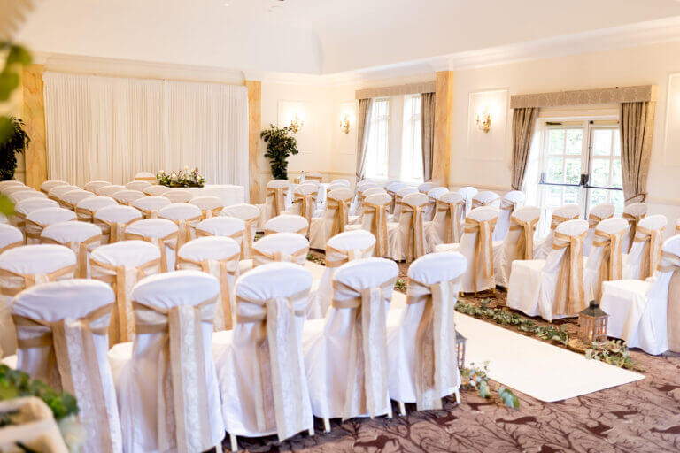 Wedding ceremony room at Careys Manor hotel with white carpet, head table and white chairs, lots of green foliage lining the walkway and lanterns with candles