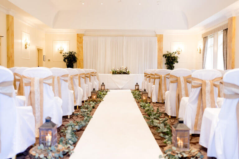 Wedding ceremony room at Careys Manor hotel with white carpet, head table and white chairs, lots of green foliage lining the walkway and lanterns with candles