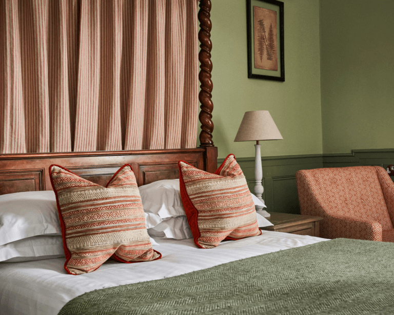 Close-up of an oak four-poster bed with striped drapes and cushions, a green wool throw and sage green walls.